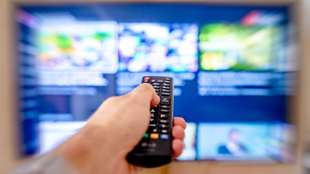 Person holding a TV remote control in front of a blurred television screen, representing VPN streaming access
